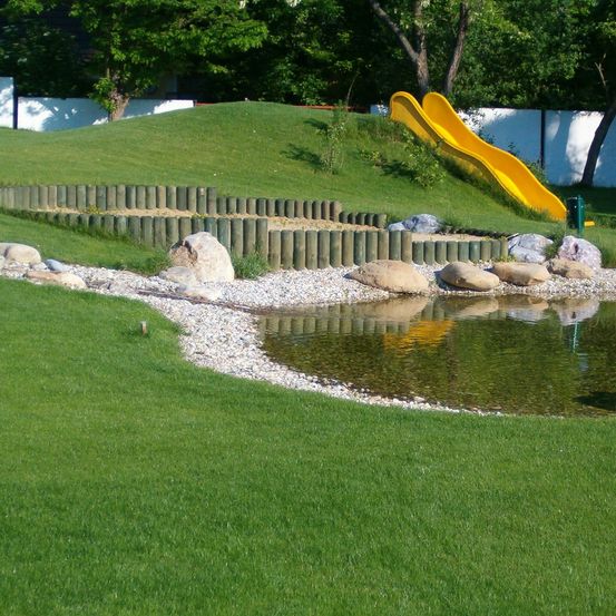 Ein Outdoor-Spielplatz mit einer gelben Rutsche auf einem grünen Hügel, umgeben von Felsen und Kieselsteinen, und einem kleinen Teich im Vordergrund.