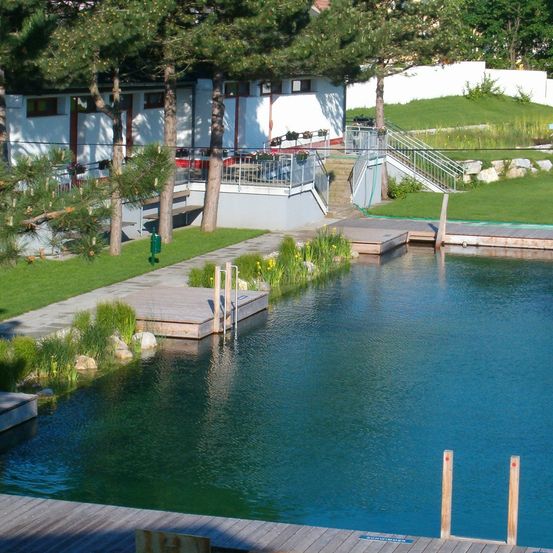 Ein großer Außenpool mit Holzdeck und Treppe ist von Bäumen und Gras umgeben. Ein weißes Gebäude mit Balkonen und Treppen ist im Hintergrund.