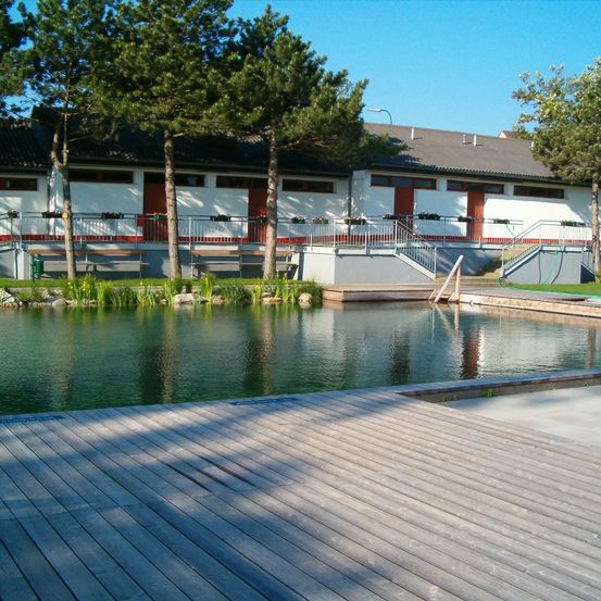 Ein ruhiger Außenbereich mit einem großen Teich, einer Holzterrasse und einem weißen Gebäude mit roten Türen und Balkonen.