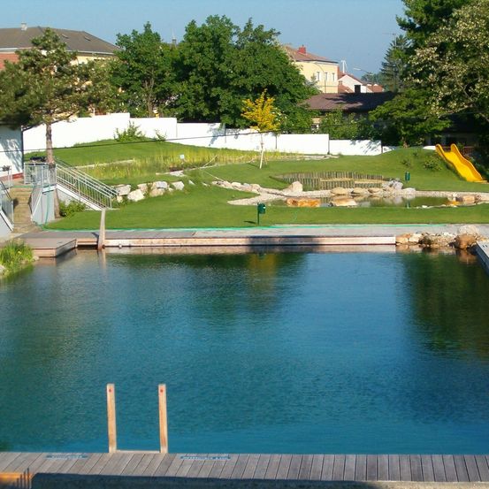 Ein ruhiger Blick auf einen Teich mit einer Holzterrasse, umgeben von einer Rasenfläche, Häusern und Bäumen. Ein gelber Rutschweg ist auf dem Grashang sichtbar.