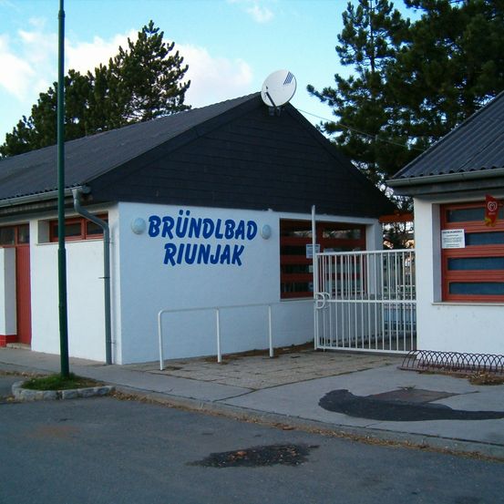 Ein Gebäude mit einem blauen Schild, das 'Brunnbad Riunjak' steht, ist zu sehen. Es hat ein weißes Tor und eine Satellitenschüssel auf dem Dach. Bäume und ein Pfosten sind im Hintergrund.