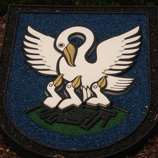 Ein Schild zeigt einen weißen Vogel mit ausgebreiteten Flügeln, ähnlich einem Schwan, auf drei kleineren Vögeln. Der Hintergrund ist blau mit einem grünen Kreis und schwarzem Text darunter.