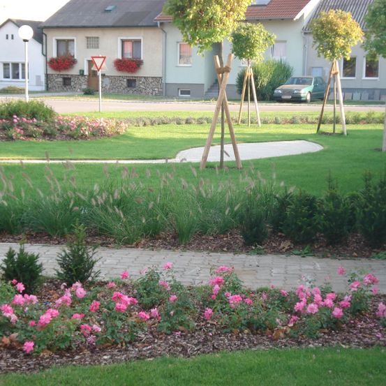Ein Park mit einem grünen Rasen und rosa Blumen, umgeben von Häusern mit roten Blumen auf den Balkonen, einem dreieckigen Schild und einem geparkten Auto.