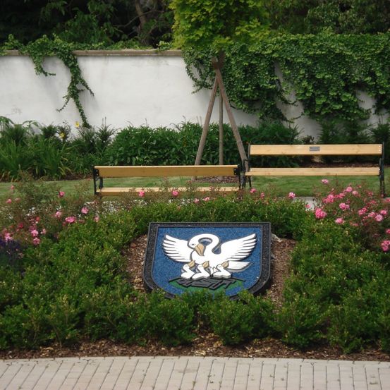 Ein Park mit zwei hölzernen Bänken und einer dekorativen Plakette mit einem weißen Vogel. Um die Plakette herum befinden sich Büsche und Blumen. Hinter den Büschen ist eine weiße Mauer teilweise mit grünen Ranken bedeckt.