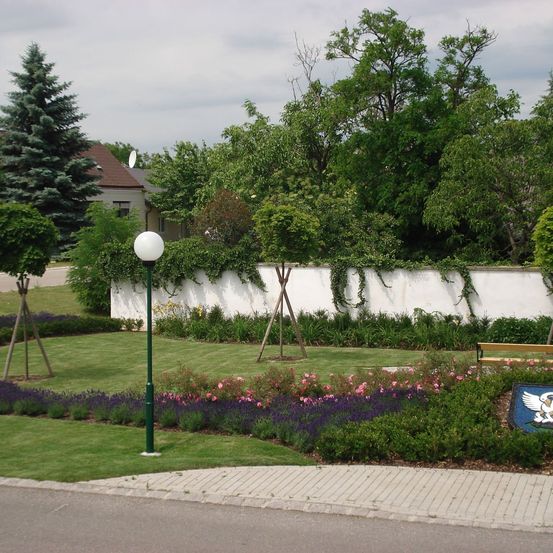 Eine friedliche Gartenszene mit einer weißen Mauer, üppigem Grün, Blumen und einer Bank. Ein Lampenpfahl steht auf dem Rasen.