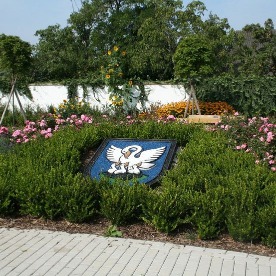 Ein Garten mit verschiedenen Blumen und Büschen, der eine blaue Plakette mit einem Schwanen-Emblem in der Mitte zeigt. Der Garten ist von Bäumen und einer weißen Mauer umgeben.