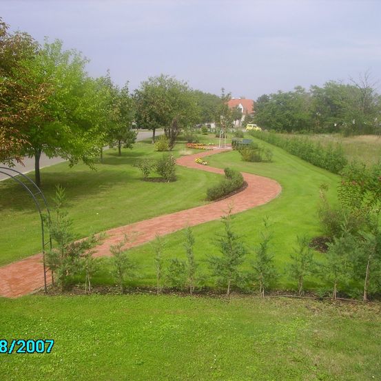 Ein gewundener Ziegelpfad durch einen grünen Park. Der Park hat Bäume, Sträucher und Blumen. Ein Haus mit einem roten Dach ist in der Ferne sichtbar. Das Foto hat ein Wasserzeichen am unteren Rand mit dem Datum 8/2007.