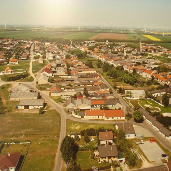 Luftaufnahme einer Stadt mit vielen Häusern, umgeben von Feldern und Windrädern in der Ferne. Die Stadt hat eine zentrale Straße mit mehreren Häusern und Gebäuden auf beiden Seiten.
