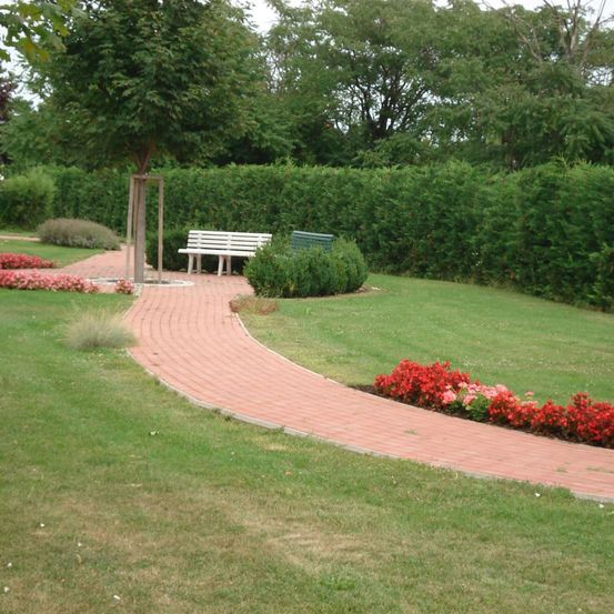 Ein gepflegter Garten mit einem Ziegelpfad, Bänken und einer Hecke. Bunte Blumen säumen den Weg, und eine Linde steht in der Mitte. Der Rasen ist ordentlich gemäht.