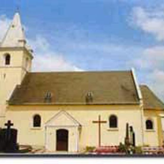 Eine alte Kirche mit einem spitzen Turm, einem braunen Dach und einem Kreuz über dem Eingang. Sie hat mehrere Rundbogenfenster und ist gelb gestrichen.