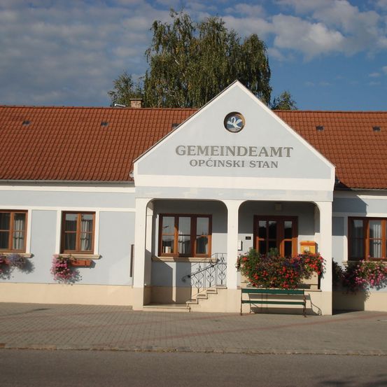 Ein weißes Gebäude mit einem roten Dach und einem Schild, das 'GEMEINDEAM OPCINSKI STAN' besagt. Es hat eine Bank davor und ist von Blumen umgeben.