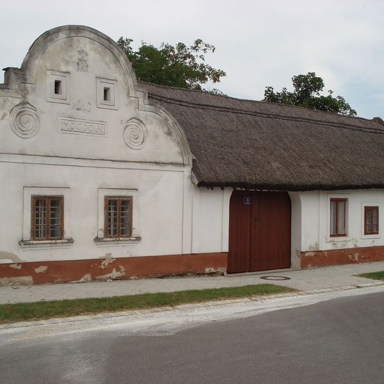 Ein altes weißes Gebäude mit einem Strohdach, einer braunen Holztür und drei Fenstern an der Vorderseite. Das Erdgeschoss ist braun gestrichen, und das Obergeschoss ist weiß.