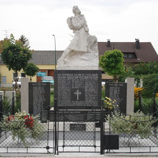 Ein Denkmal in einem Freiluftfriedhof zeigt eine Statue einer Frau mit einem Baby, umgeben von einem Zaun. Der Sockel hat Tafeln mit Namen und Daten. Dahinter ist ein Gebäude mit einem braunen Dach zu sehen.