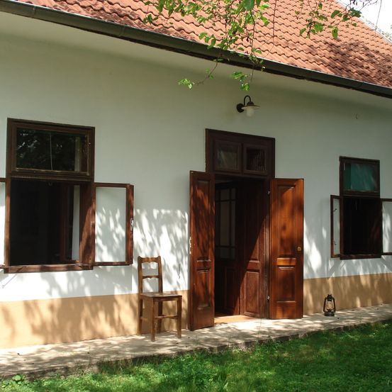 Ein weißes Haus mit einem roten Dach hat eine offene Tür und Fenster. Ein Holzstuhl steht auf dem Vordach.