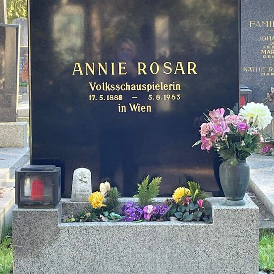 Ein Granitgrabstein mit schwarzem Hintergrund zeigt den Namen Annie Rosar, eine Volksschauspielerin, geboren am 17. Mai 1888 und gestorben am 5. August 1963. Ein Blumentopf mit Blumen und eine Kerze stehen in einem kleinen Pflanzgefäß davor.