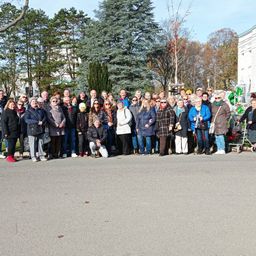 Eine große Gruppe von Erwachsenen ist auf einer Straße versammelt, lächelt und posiert für ein Foto. Einige tragen Brillen und Hüte. Bäume und ein Gebäude sind im Hintergrund zu sehen.
