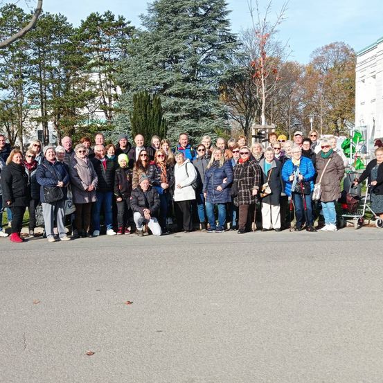 Eine große Gruppe von Erwachsenen ist auf einer Straße versammelt, lächelt und posiert für ein Foto. Einige tragen Brillen und Hüte. Bäume und ein Gebäude sind im Hintergrund zu sehen.