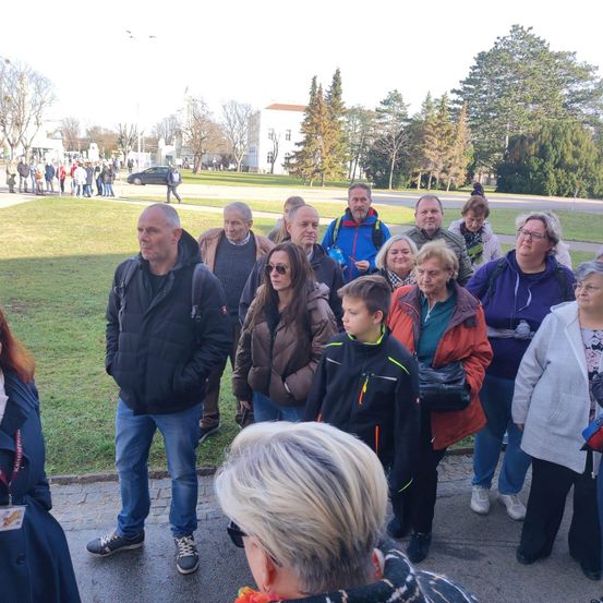 Eine Gruppe von Menschen, darunter Erwachsene und Kinder, versammelt sich im Freien. Sie tragen alle Jacken, einige von ihnen tragen Brillen. Die Frau in der Mitte trägt eine Sonnenbrille. Bäume und ein Gebäude sind in der Ferne zu sehen.