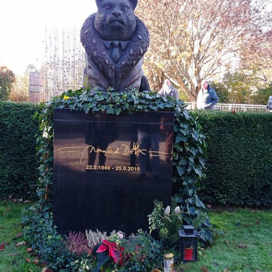 Eine Statue eines Bären in Pelzmantel und Krawatte steht auf einem schwarzen Podest. Efeu und Blumen umgeben die Basis. Dahinter stehen Menschen in der Nähe eines Zauns.
