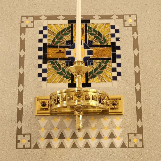 Ein goldener Kerzenhalter mit einer weißen Kerze ist an einer Wand mit geometrischem Muster montiert. Die Kerze ist von einem farbenfrohen Quadratdesign umgeben.