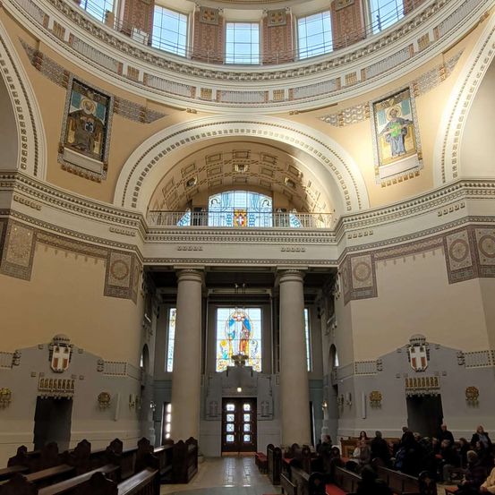Ein Innenraum einer großen Kathedrale mit hoher gewölbter Decke, Buntglasfenstern und aufwendigen Kunstwerken. Säulen säumen den Mittelgang, der zu einem zentralen Altar führt. Die Menschen sitzen auf hölzernen Kirchenbänken.