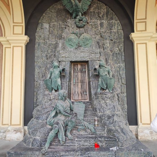 Ein Steinmonument mit einer sitzenden Figur, die ein Buch hält, flankiert von Statuen zweier Wachen mit Fackeln und einer Holztür mit einem Kranz darüber.
