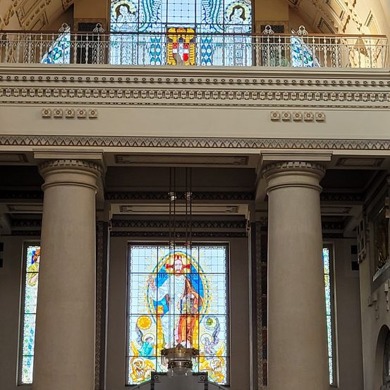 Das Innere einer großen Kirche mit großen Buntglasfenstern und einer prominenten zentralen Figur. Hohe Säulen stützen die Struktur, mit dekorativen Geländern darüber.
