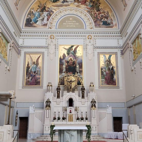 Ein Altar in einer Kirche mit religiösen Gemälden an den Wänden. Der Altar hat Blumen und Kerzen. Die Decke hat ein kompliziertes Design mit einem goldenen Rahmen.