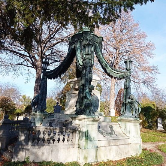 Ein großes, kunstvoll verziertes Denkmal mit Statuen steht auf einem Friedhof. Es hat ein Baldachin mit dekorativen Elementen und eine Steinbalustrade. Die Statuen umfassen eine Figur mit Schild und eine Lampe.