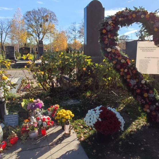 Ein herzförmiger Blumenkranz schmückt einen Friedhof. Es gibt mehrere Grabsteine und Blumensträuße. Im Hintergrund stehen Bäume mit gelben und orangefarbenen Blättern. Ein Schild mit deutschem Text ist an der Wand befestigt.