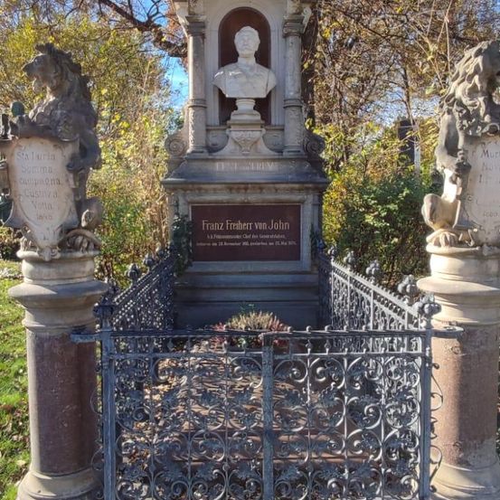 Ein Denkmal mit einer Büste von Franz Freiherr von John, umgeben von zwei Löwenstatuen, ist von einem eisernen Zaun eingeschlossen. Das Denkmal befindet sich in einem Grasbereich mit Bäumen.
