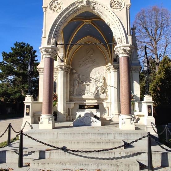 Ein großes, kunstvolles Marmordenkmal mit einem Spitzbogen, zwei Säulen und einer komplexen Reliefskulptur. Das Denkmal ist von einer Kettenumzäunung auf einer Betonplattform umgeben. Bäume und Pflanzen sind im Hintergrund sichtbar.
