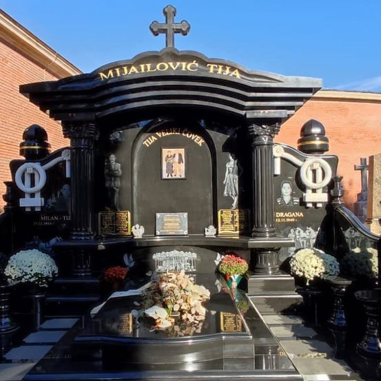 Ein schwarzer Grabstein mit einem Kreuz und dem Namen Mijailovic TiJa. Er hat mehrere Plaketten und einen Fotorahmen, zusammen mit frischen Blumen. Dahinter steht ein Gebäude mit Ziegelwänden.