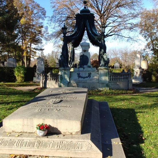 Ein Grabstein mit einer Statue und einem Baldachin auf einem Friedhof, umgeben von Bäumen und Gras, mit einer kleinen Blumengestecke darauf.