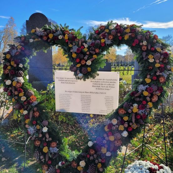 Ein herzförmiger Kranz aus Blumen und Tannenzapfen liegt auf einem Grabstein. Eine weiße Tafel mit Text und Namen befindet sich in der Mitte. Es ist vor einem Hintergrund aus Bäumen und Gras platziert.