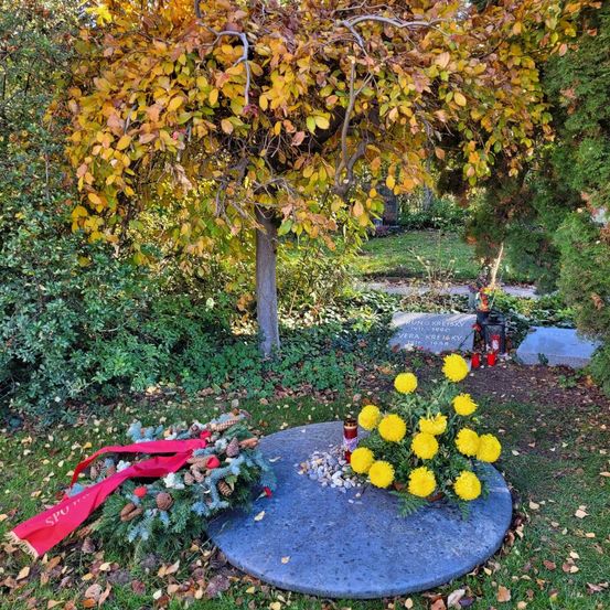 Ein Denkmal auf einem Friedhof besteht aus einer runden Steinplatte mit einer gelben Blumenanordnung, umgeben von einem Kranz und einem roten Band, mit einem Grabstein und einer Laterne im Hintergrund.