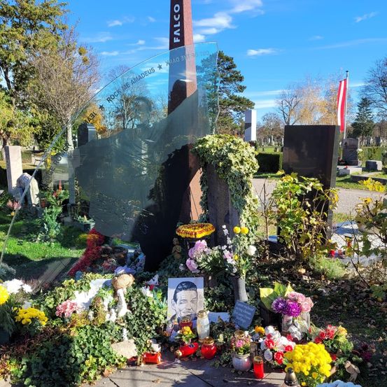 Ein Denkmal auf einem Friedhof mit einer großen Glasplakette, die eine Silhouette einer Person zeigt, umgeben von Blumen, Pflanzen und einem Bilderrahmen. Ein Pfahl mit dem Wort Falco ist sichtbar, und eine Flagge befindet sich rechts.