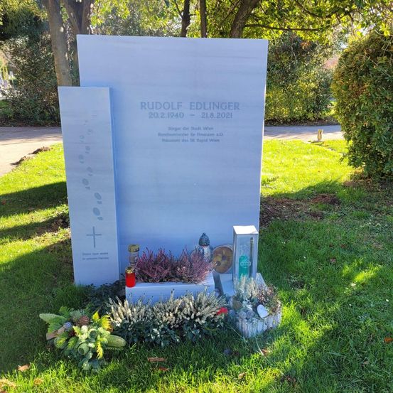 Ein Gedenkstein für Rudolf Edlinger, geboren am 20. Februar 1940 und verstorben am 21. August 2021, steht auf einer Rasenfläche. Er beinhaltet ein Kreuz, Blumen und Kerzen.