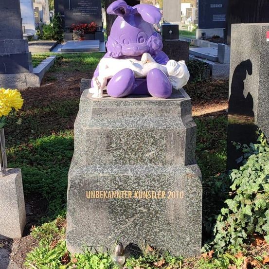 Eine lila Statue eines Osterhasen sitzt auf einem Grabstein. Der Stein trägt die Aufschrift 'Unbekannter Künstler 2010'. Das Grab befindet sich auf einem Friedhof, in der Nähe einer gelben Blume.