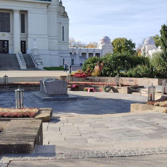 Ein breiter gepflasterter Bereich vor einem großen weißen Gebäude mit einem Gedenkstein, umgeben von Büschen und Blumen. Eine Person geht auf dem Rasen im Hintergrund.