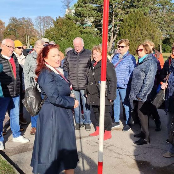 Eine Gruppe von Menschen in Winterkleidung steht in einer Reihe neben einem rot-weißen Pfosten. Bäume und ein Gebäude sind im Hintergrund zu sehen.