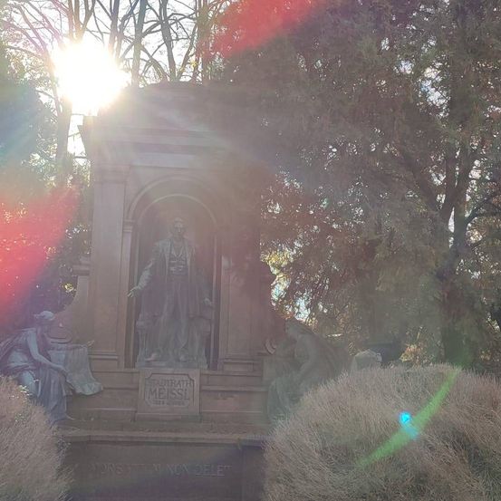Eine Statue von Friedrich von Flotow steht unter einem hellen Sonnenschein, umgeben von zwei liegenden Figuren und Bäumen.