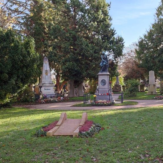 Ein Friedhof mit einem Denkmal in der Mitte, umgeben von üppigem grünen Bäumen und Pflanzen. Das Denkmal hat eine Statue einer sitzenden Person. Der Bereich ist von einem Zaun mit Blumen umgeben.