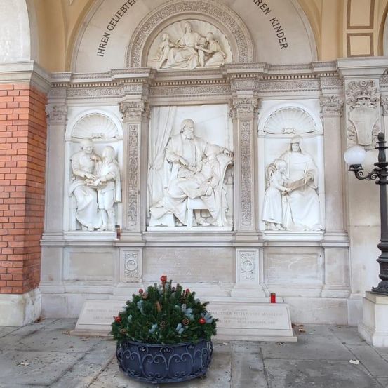 Ein Denkmal mit Statuen, die geliebten Kindern gewidmet ist. Die zentrale Figur zeigt einen sitzenden Mann mit einem Kind, flankiert von zwei weiteren Statuen. Oben zeigt ein Relief eine sitzende Figur mit Kindern. Vorne steht ein kleiner immergrüner Kranz.
