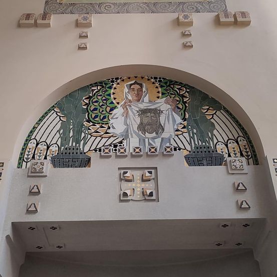 Ein Mosaik-Wandbild an einem Bogen zeigt eine Frau, die ein Tuch mit einer bärtigen Figur hält. Geometrische Muster schmücken den Bogen und die Wand.