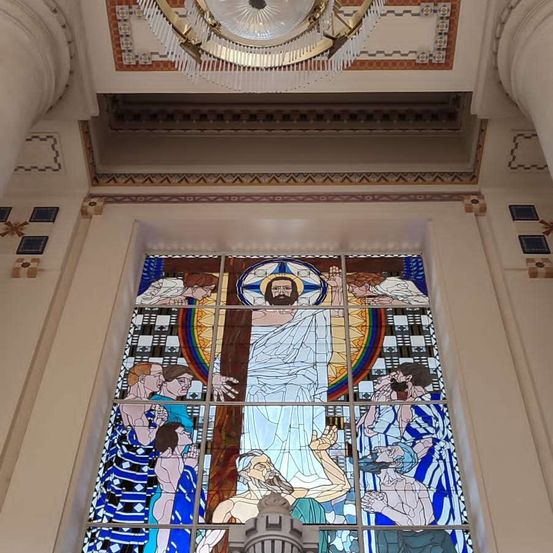 Ein Buntglasfenster zeigt Jesus mit einem Regenbogen und Menschen in verschiedenen Szenen. Die Wand hat geometrische Muster.