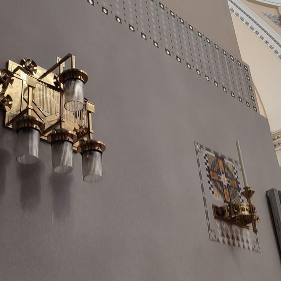 Eine goldene Wandlampe mit drei zylindrischen Glasschirmen ist an einer grauen Wand befestigt. Auf der rechten Seite befindet sich eine dekorative Wandfliese mit einem weißen Kerzenhalter.