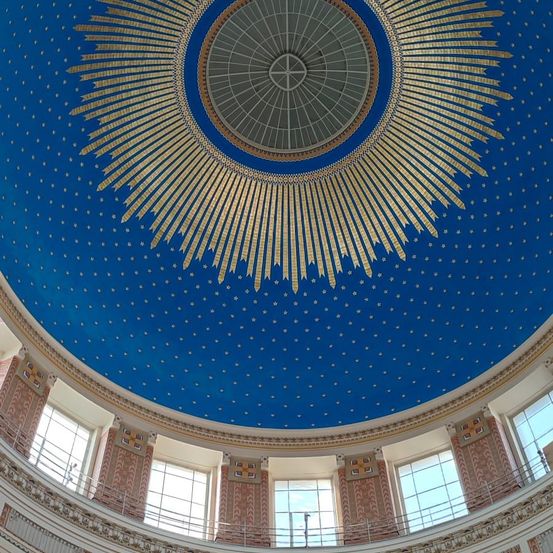 Eine runde blaue Kuppel mit einem goldenen Sonnenstrahlmuster in der Mitte, umgeben von zahlreichen Sternen. Die Kuppel wird von einer Reihe von Fenstern und dekorativen Bögen gesäumt.