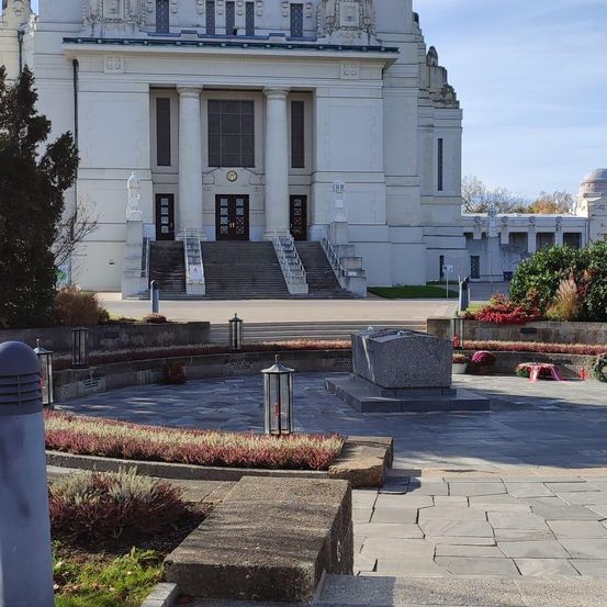 Ein großes weißes Gebäude mit klassischer Architektur, mit großen Säulen und einem Kuppeldach. Stufen führen zum Eingang, umgeben von einem Rasen mit verstreuten Blumen. Ein grauer Steinmonument steht im Vordergrund.