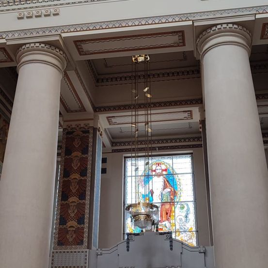 Ein großer Innenraum mit hohen Säulen, einer Kronleuchter und einem Buntglasfenster, das eine religiöse Figur darstellt.